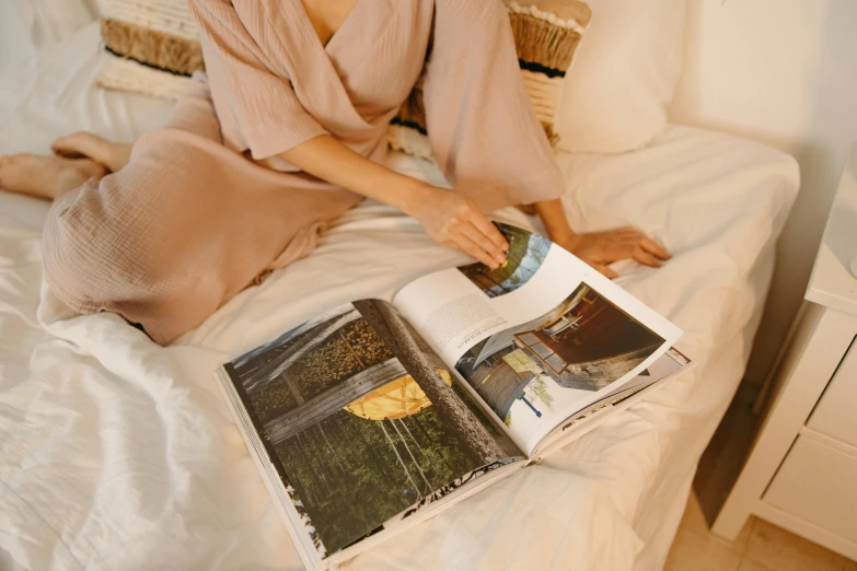 a woman sitting on a bed reading a book, a picture, by Carey Morris, pexels contest winner, earthy colours, magazine pages, wearing simple robes, bottom angle