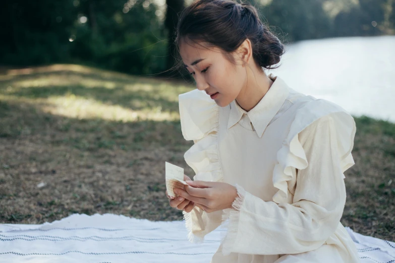 a woman sitting on a blanket looking at her cell phone, inspired by Cui Bai, pexels contest winner, paper origami, victorian inspired clothing, wearing a linen shirt, sydney park