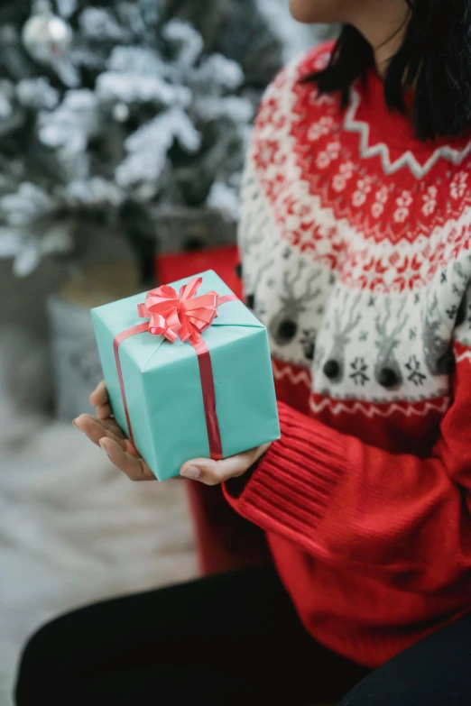 a woman sitting in front of a christmas tree holding a present, by Julia Pishtar, pexels contest winner, red and teal color scheme, sweater, opening shot, gif