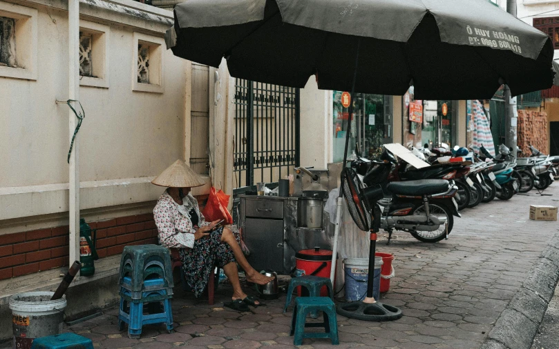 a woman sitting under an umbrella on a sidewalk, with street food stalls, hoang long ly, profile image, high resolution photo