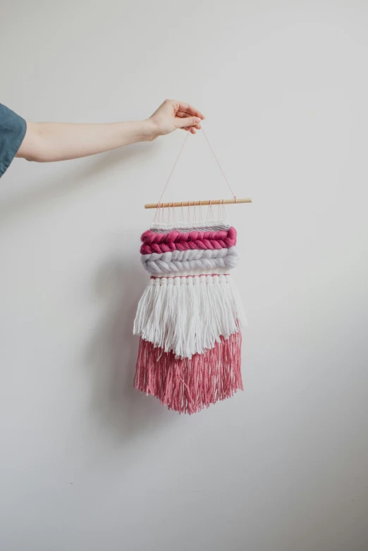 a person holding a yarn hanging on a wall, white and pink, full product shot, loom, reds)