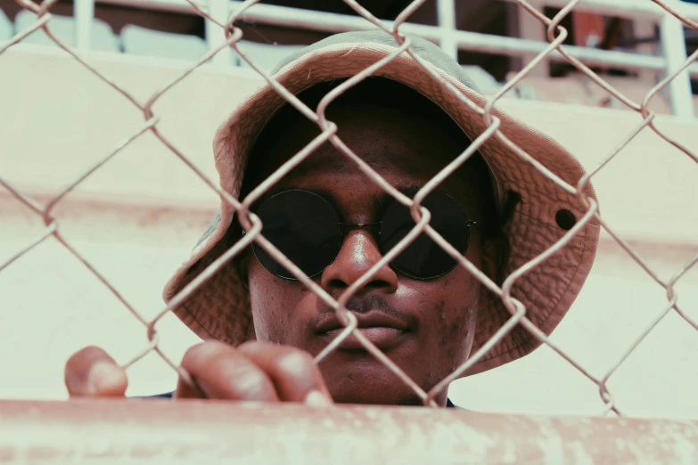 a man wearing a hat and sunglasses behind a fence, a picture, pexels contest winner, afrofuturism, brown skin, sunfaded, syd, bladee from drain gang
