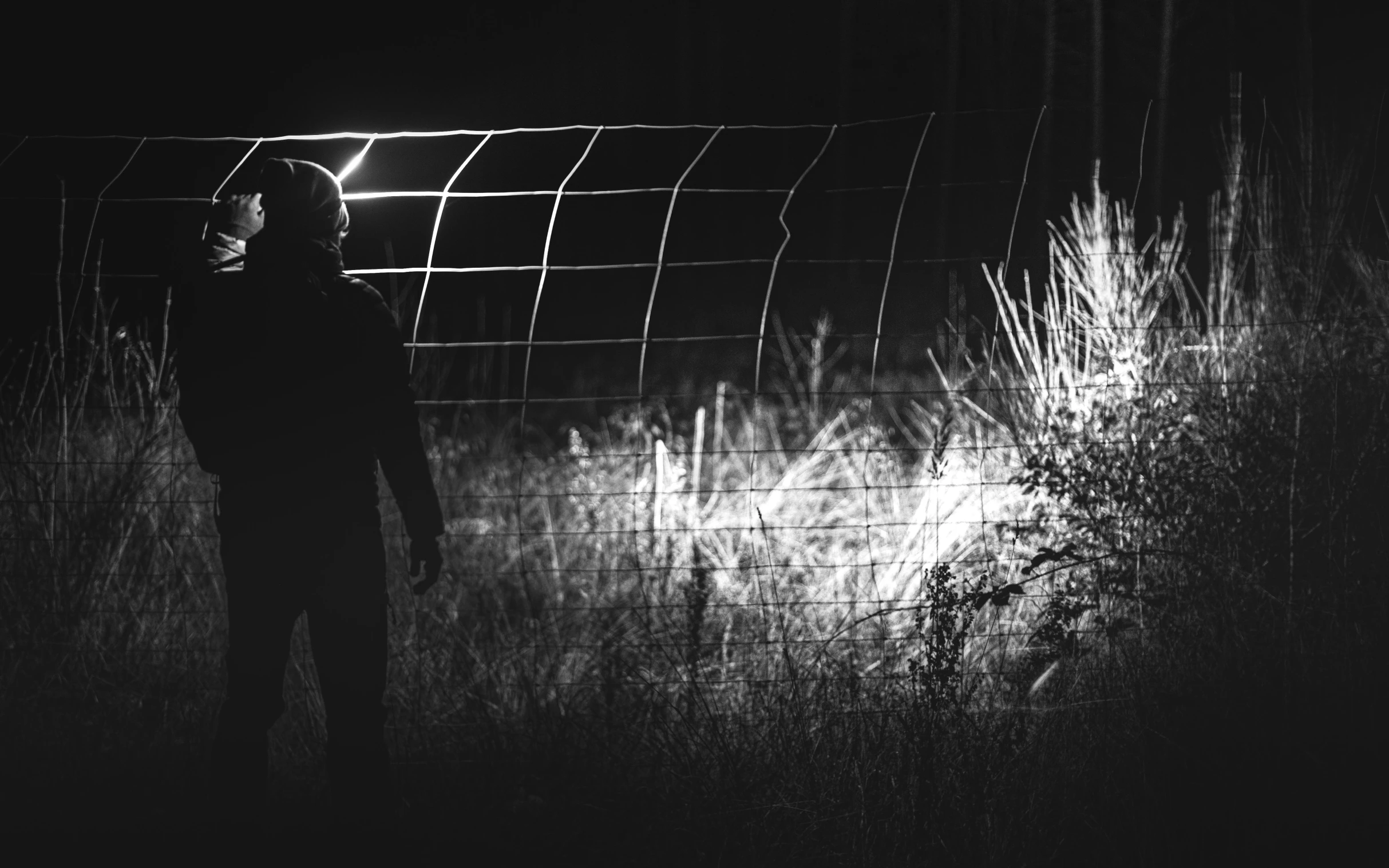 a person standing in front of a fence at night, a black and white photo, by Adam Chmielowski, unsplash, conceptual art, hunting, wire, photorealistic logan movie still, ❤🔥🍄🌪