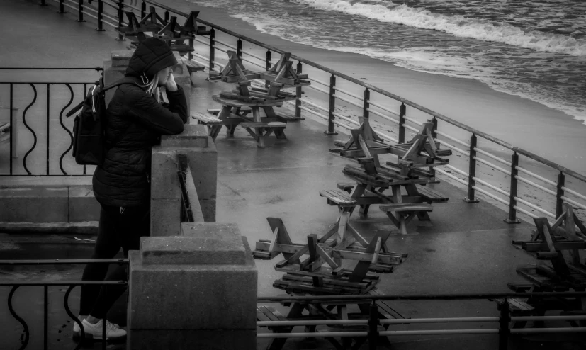 a man standing on top of a pier next to the ocean, a black and white photo, inspired by Louis Stettner, pexels contest winner, realism, people sitting at tables, cold stormy wind, benches, distant hooded figures