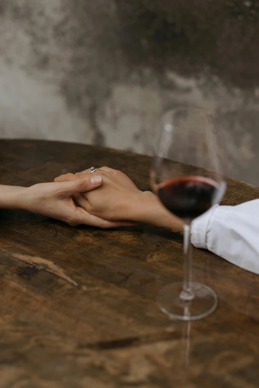 a close up of a person holding a glass of wine, holding each other hands, sitting on a table, secret romance, ignant