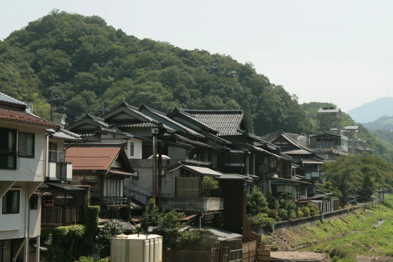 a train traveling down a train track next to a lush green hillside, inspired by Maruyama Ōkyo, sōsaku hanga, stacked houses, view from the street, houses on stilts, tiled roofs