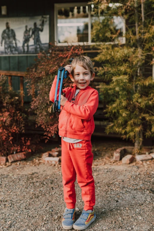 a little boy standing in front of a house, wearing a track suit, wearing adventuring gear, bright red, holding wood saw