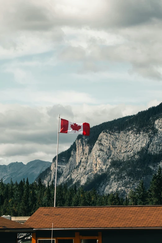 a building with a flag flying in front of a mountain, pexels contest winner, fine art, canada, red banners, hill with trees, bakelite rocky mountains