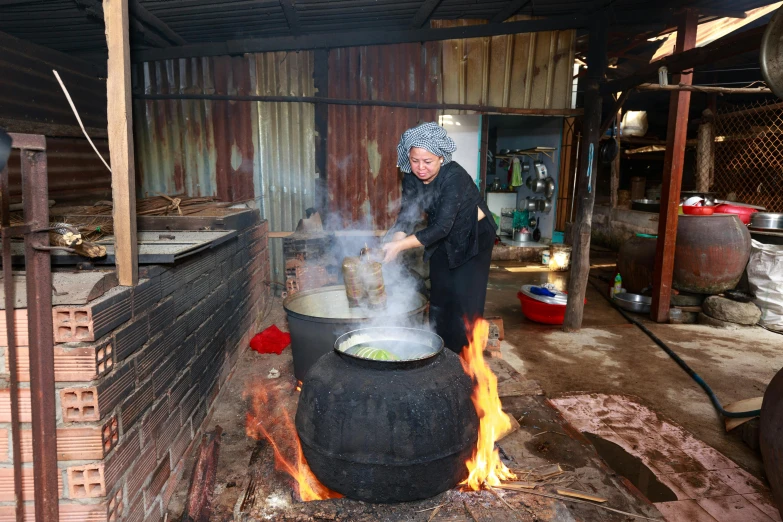 a woman cooking food in a large pot on a fire, ngai victo, profile image, thumbnail, production photo