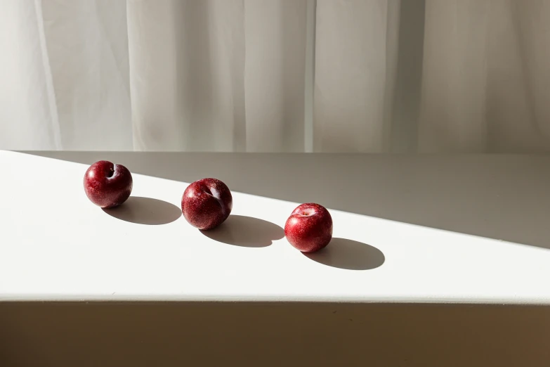 three red apples sitting on top of a white table, a still life, inspired by Robert Mapplethorpe, unsplash, sun puddle, light from right, ignant, in a row