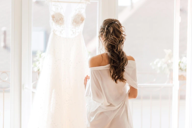 a woman standing in front of a window looking at a wedding dress, pexels contest winner, happening, wearing a white hospital gown, on a white table, looking from behind, looking cute