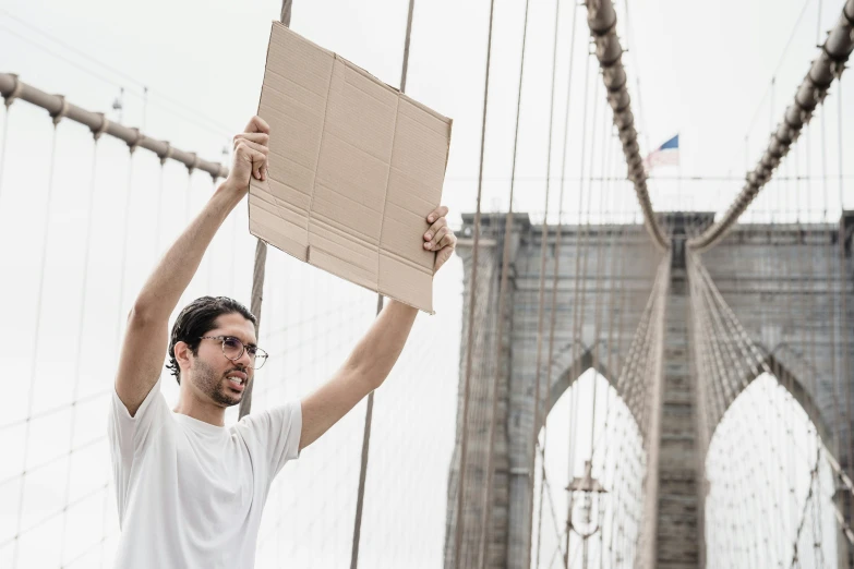 a man holding up a box on top of a bridge, inspired by Christo, pexels contest winner, protesters holding placards, brooklyn, made of cardboard, promotional image