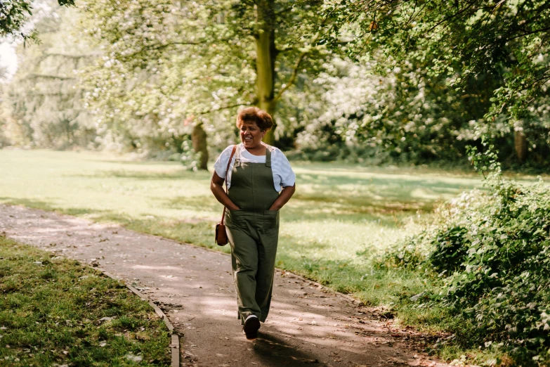 a woman walking down a path in a park, by Helen Stevenson, happening, wearing overalls, healthy, press shot, green spaces