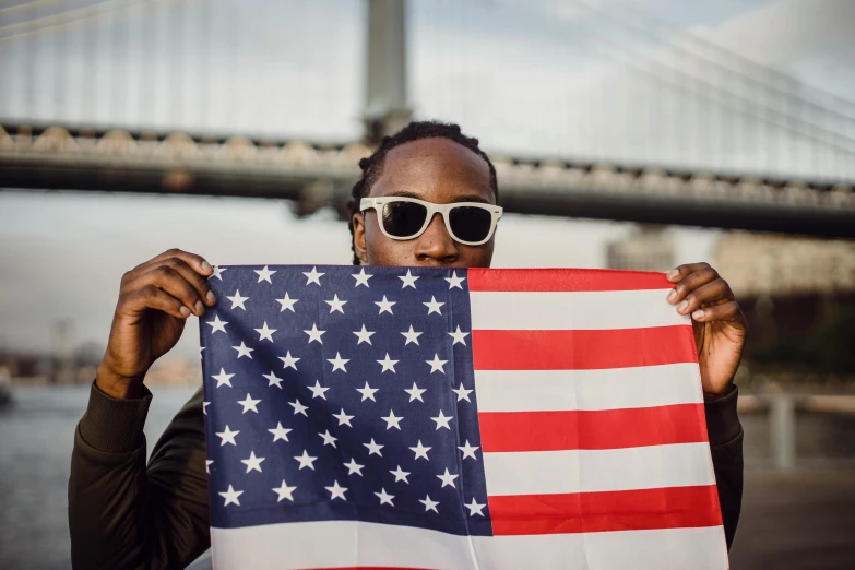 a man holding an american flag in front of a bridge, pexels contest winner, young thug, shades, 🚿🗝📝, lebron james solemn at tea party