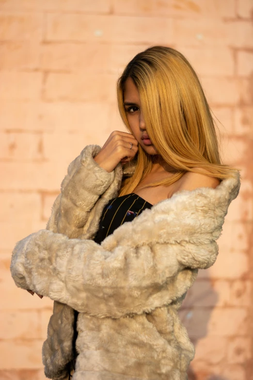 a woman standing in front of a brick wall, an album cover, by Robbie Trevino, trending on pexels, renaissance, fur jacket, light gold hair, mia khalifa, detail shot