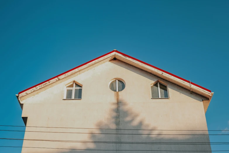 the shadow of a tree on the side of a building, by Adam Marczyński, pexels contest winner, simple gable roofs, clear blue sky, round windows, suburban home