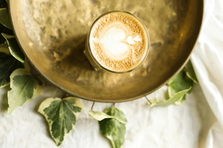 a cup of coffee sitting on top of a table, by Emma Andijewska, trending on unsplash, renaissance, moringa juice, white and gold color palette, ivy, vanilla smoothie explosion