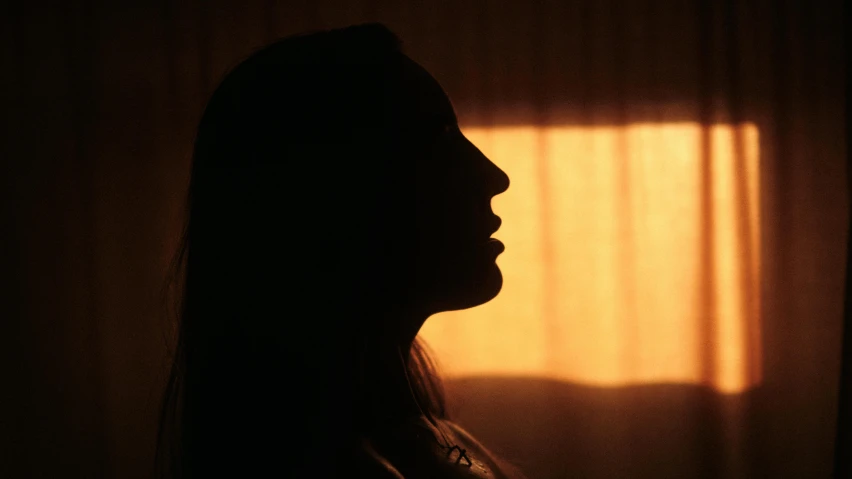 a silhouette of a woman standing in front of a window, a picture, orange light, side profile shot, close-up portrait film still, low angle photograph
