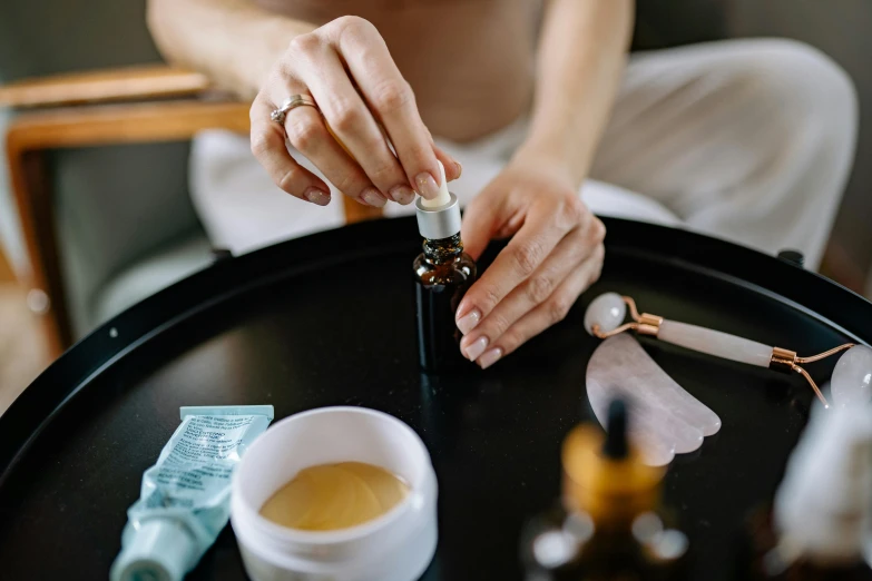 a woman sitting at a table with a bottle of essential oils, by Julia Pishtar, trending on pexels, putting makeup on, manuka, candy treatments, thumbnail