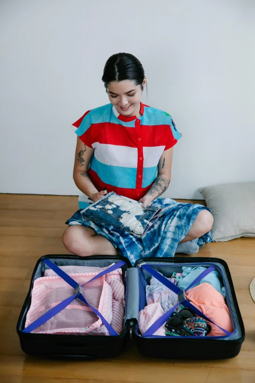 a woman sitting on the floor with a suitcase full of clothes, wearing a tie-dye shirt, tattooed, in suitcase, guide