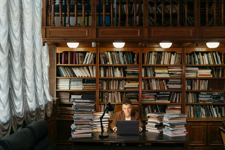 a man sitting at a desk in front of a laptop computer, by Julia Pishtar, pexels contest winner, academic art, bookshelves on sides, stack of books on side table, sydney hanson, inside a grand