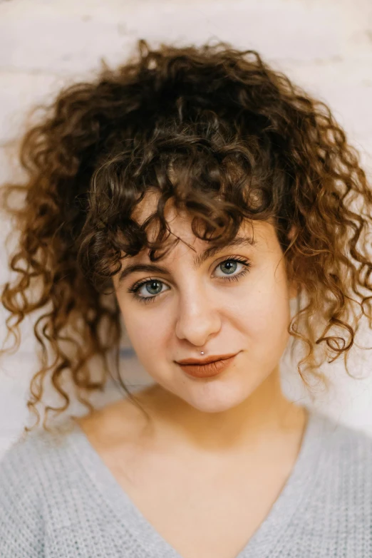 a woman with curly hair standing in front of a brick wall, inspired by Elizabeth Polunin, closeup headshot, actress, rebecca sugar, on grey background