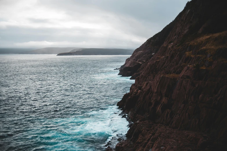 a person standing on top of a cliff next to the ocean, by Carey Morris, pexels contest winner, “ iron bark, cut into the side of a mountain, brown, viewed from bellow