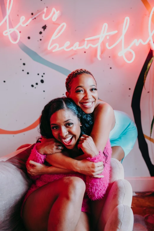 two women sitting on a couch in front of a neon sign, a portrait, by Dulah Marie Evans, trending on pexels, vanessa morgan, hugging each other, both smiling for the camera, promotional image