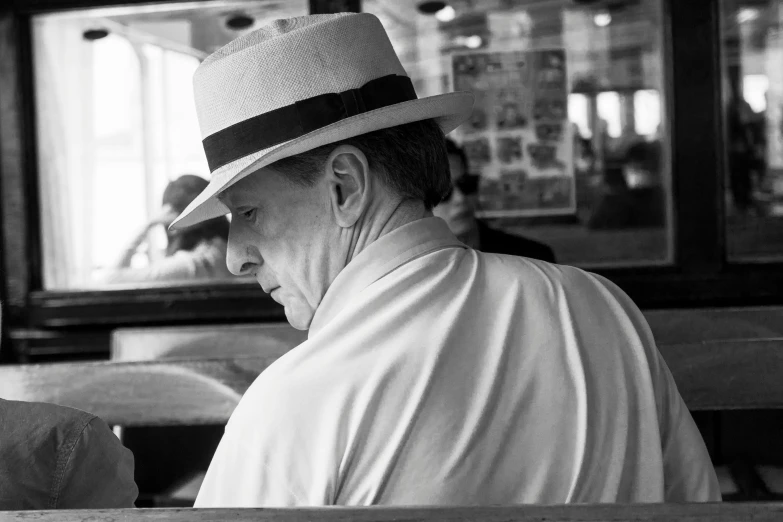 a black and white photo of a man sitting on a bench, inspired by Vivian Maier, white straw flat brimmed hat, in classic diner, profile image, wearing white shirt