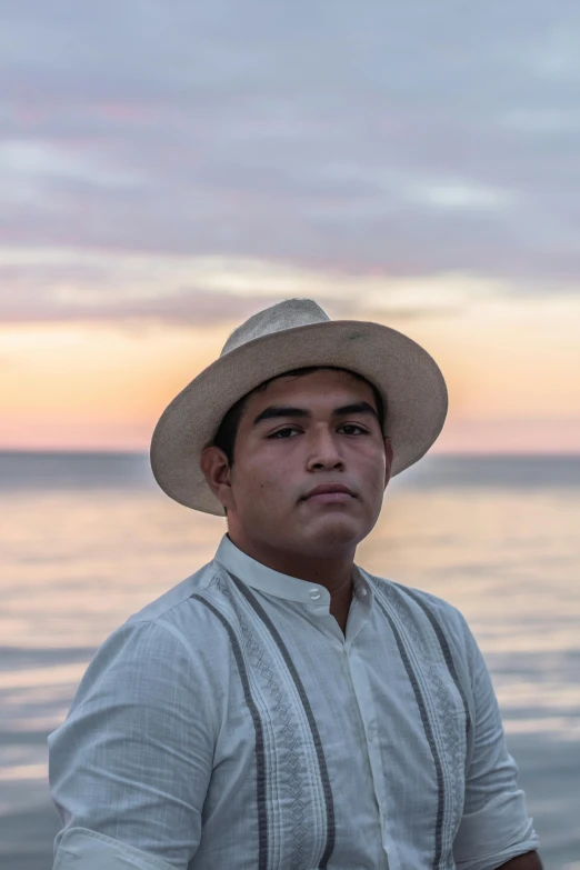 a man in a hat sitting next to a body of water, a portrait, inspired by Rodolfo Morales, unsplash, folklorico, sunset in background, avatar image, wearing a linen shirt