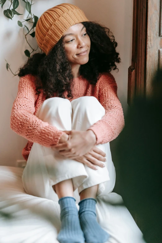 a woman sitting on top of a bed next to a plant, inspired by Anita Malfatti, trending on pexels, renaissance, knitted hat, mixed race woman, orange hoodie, full body close-up shot