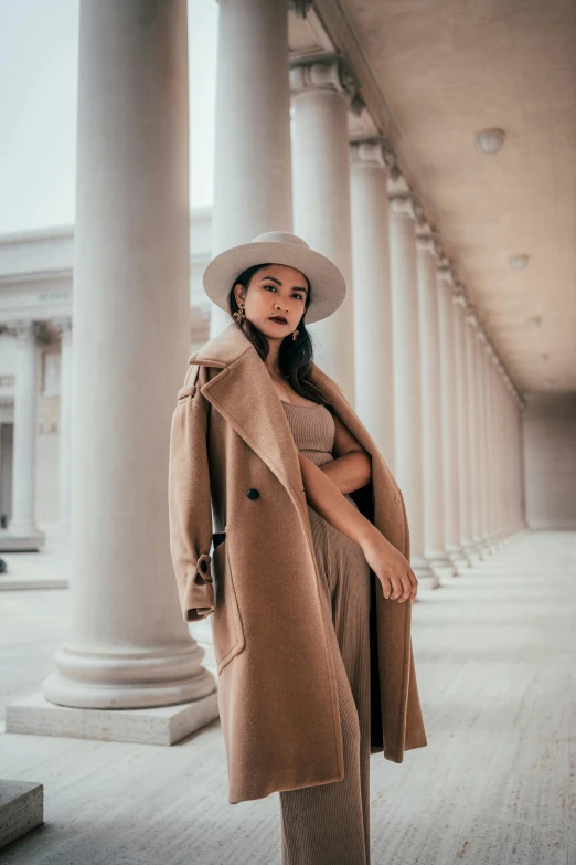 a woman standing in front of a building wearing a coat and hat, trending on pexels, renaissance, malaysian, wearing long gown, tan, bella poarch