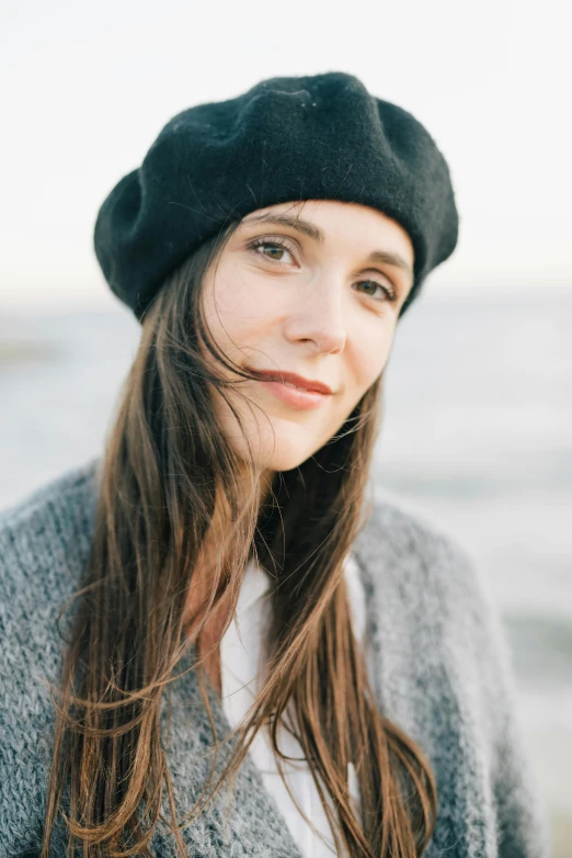 a woman standing on top of a beach next to the ocean, a portrait, inspired by Anita Malfatti, unsplash, wearing a beret, girl with dark brown hair, headshot, soft oval face