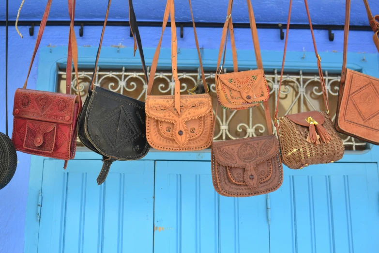 a bunch of purses hanging from a blue door, a photo, by Hannah Tompkins, trending on unsplash, arabesque, intricate leather suspenders, square, moroccan, blue sky