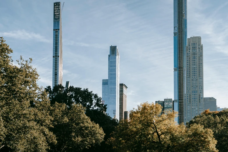 a group of people sitting on top of a lush green field, inspired by Thomas Struth, unsplash contest winner, visual art, new york buildings, three towers, during autumn, with dark trees in foreground