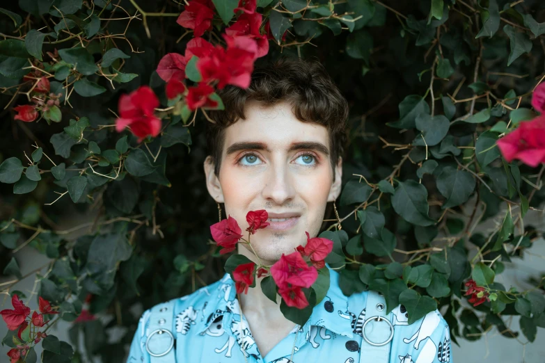 a man standing in front of a bush of flowers, an album cover, by Julia Pishtar, pexels contest winner, androgynous face, asmongold, light stubble with red shirt, made of flowers and leaves