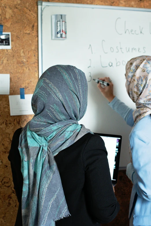 two women are writing on a white board, hurufiyya, coding, costume desig, wearing a head scarf, production photo