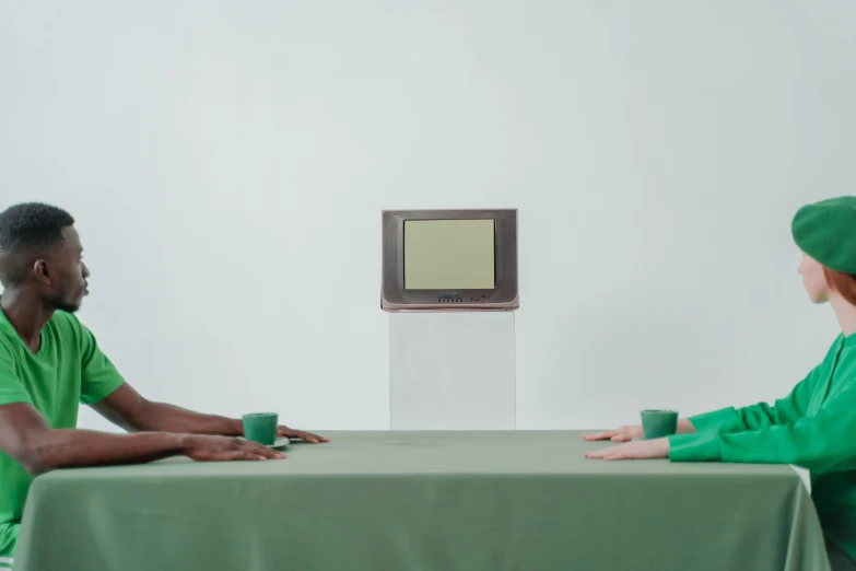 two men sitting at a table in front of a television, inspired by Vanessa Beecroft, unsplash, video art, green and brown clothes, on a pale background, four hands, with people watching in wonder