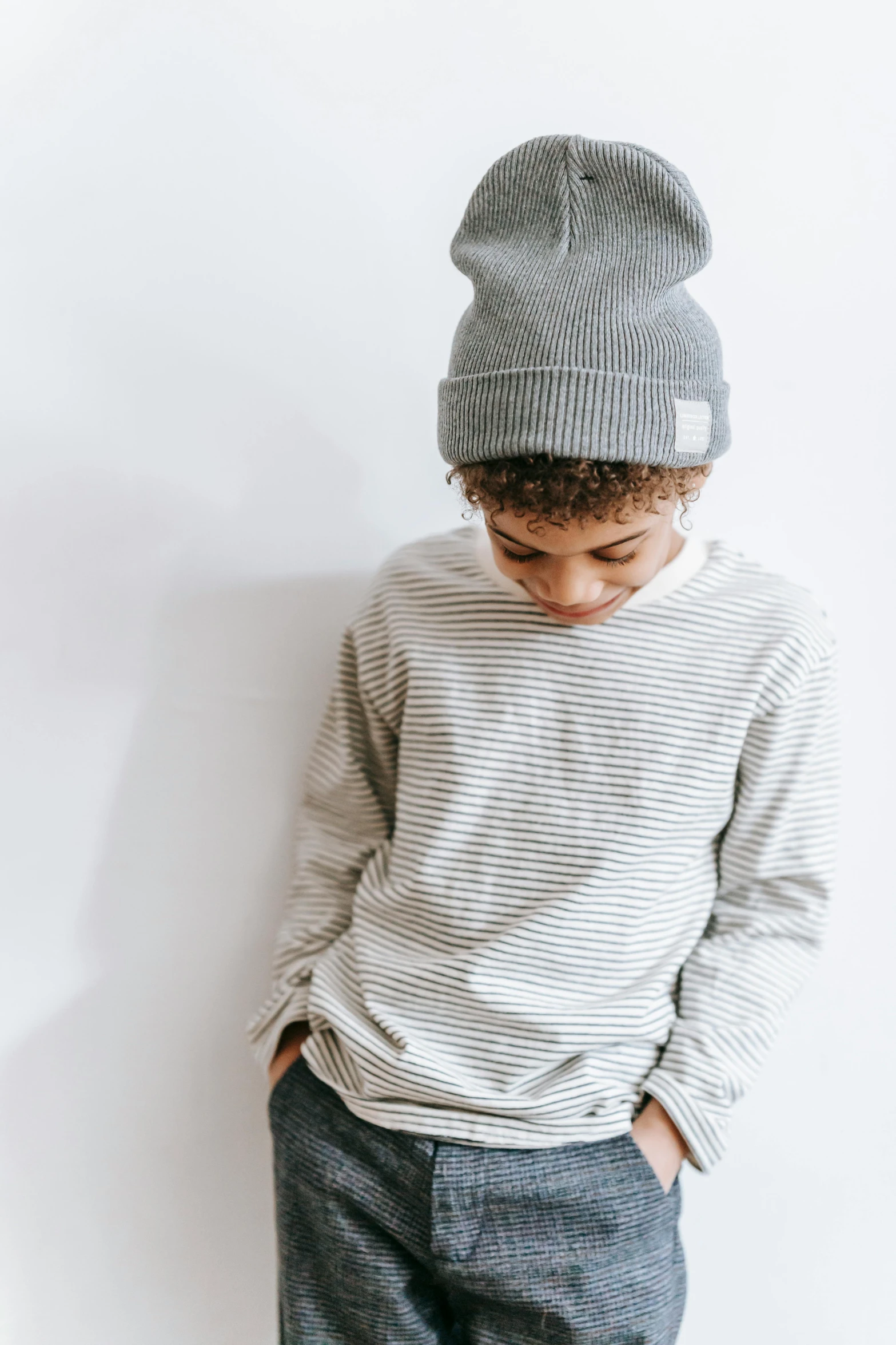 a young boy standing in front of a white wall, inspired by Knox Martin, trending on unsplash, beanie, light grey, striped shirt, long sleeves