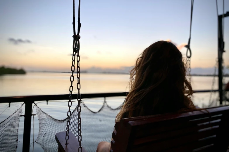 a woman sitting on a swing overlooking a body of water, profile image, on the deck of a ship, sun down, tourist photo