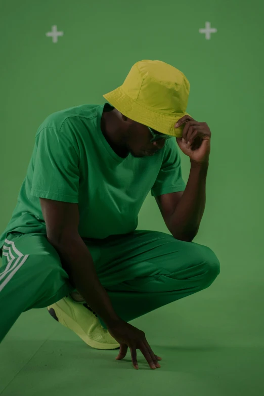 a man squatting in front of a green screen, an album cover, trending on unsplash, visual art, yellow cap, wearing adidas clothing, directional sunlight skewed shot, bucket hat