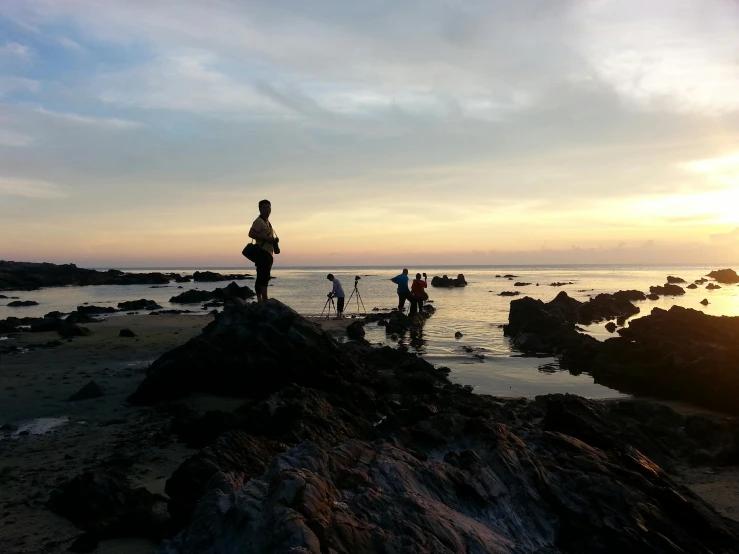 a group of people standing on top of a rocky beach, in the sunset, thawan duchanee, rock pools, fishing