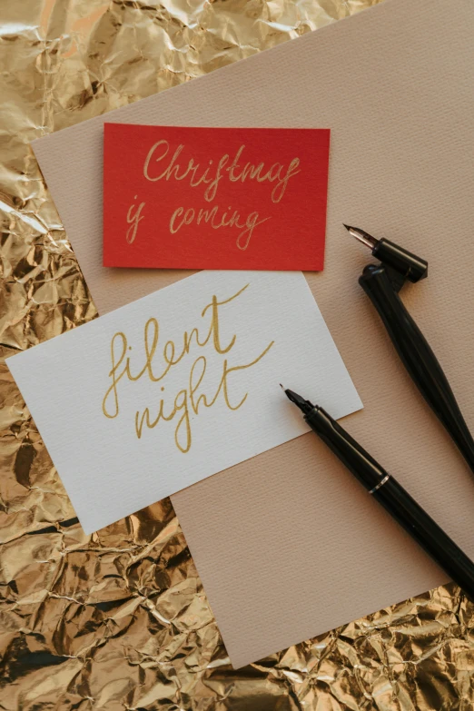 a couple of pens sitting on top of a piece of paper, inspired by Christian Hilfgott Brand, happening, festive atmosphere, tin foiling, with some hand written letters, shining gold and black and red