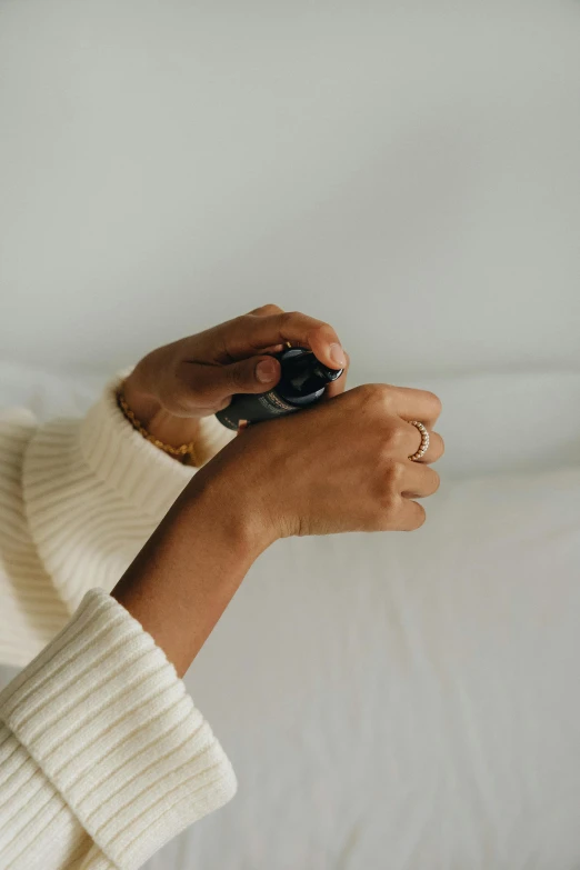 a close up of a person holding a camera, perfume bottle, white bed, wearing casual sweater, shungite bangle
