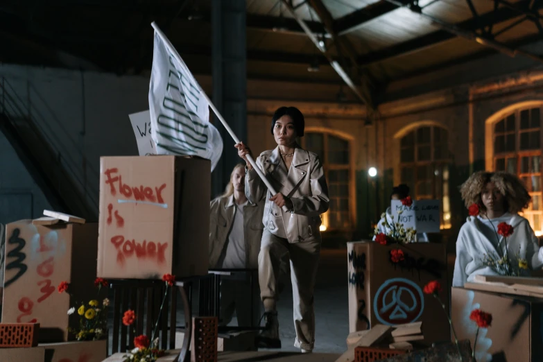 a woman holding a flag in a room full of boxes, inspired by Fei Danxu, carrying flowers, brutalist fashion show, protesters holding placards, on set