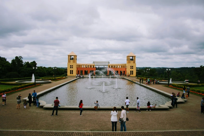 a group of people standing around a fountain, inspired by Maties Palau Ferré, pexels contest winner, building in the distance, crystal palace, colombia, on a cloudy day