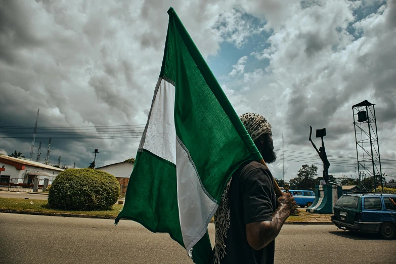 a man holding a large green and white flag, an album cover, by Chinwe Chukwuogo-Roy, unsplash, symbolism, neighborhood, independence, tear gas, a pilgrim