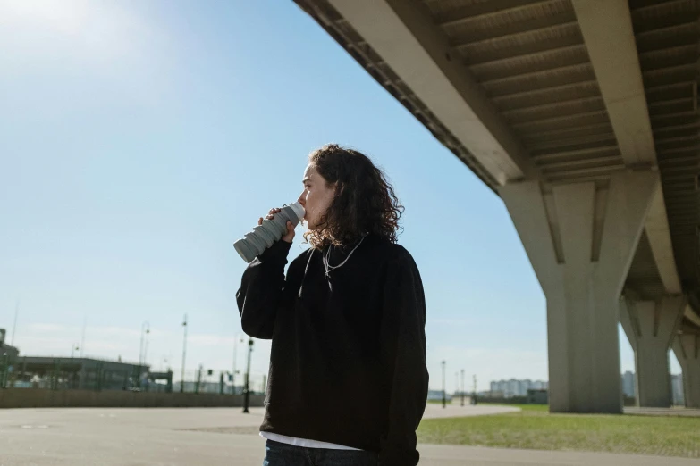 a man standing under a bridge drinking from a cup, unsplash, happening, black haired girl wearing hoodie, on a sunny day, profile image, vapourwave