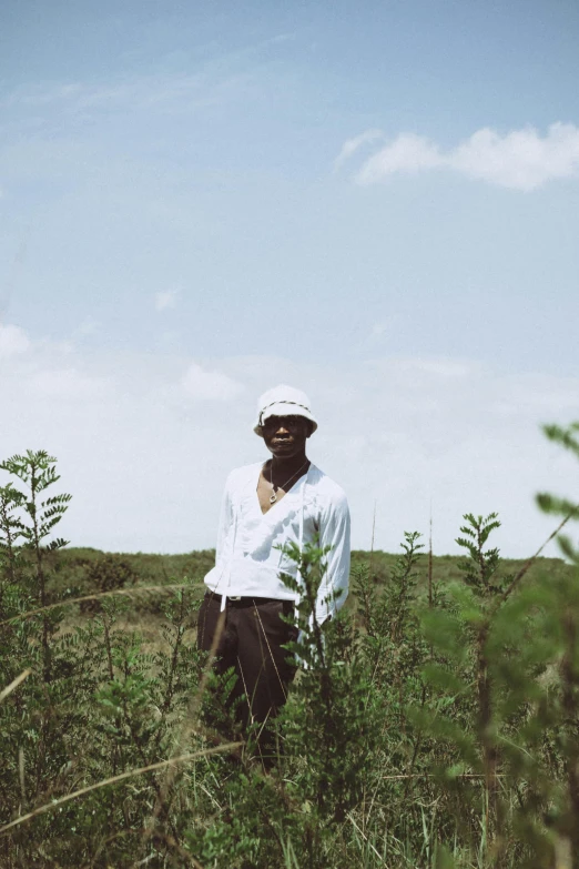 a man standing in a field of tall grass, an album cover, inspired by Barthélemy Menn, unsplash, wearing white clothes, he is wearing a hat, dark skinned, sea - green and white clothes