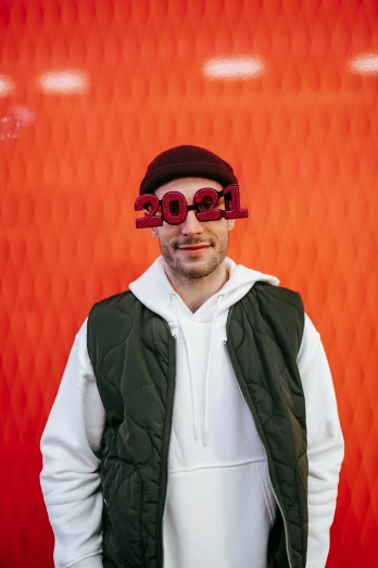 a man standing in front of a red wall, an album cover, inspired by Josh Bayer, trending on pexels, futurism, goggles on forehead, he is wearing a hat, 2 0 2 2 picture of the year, charlie cox
